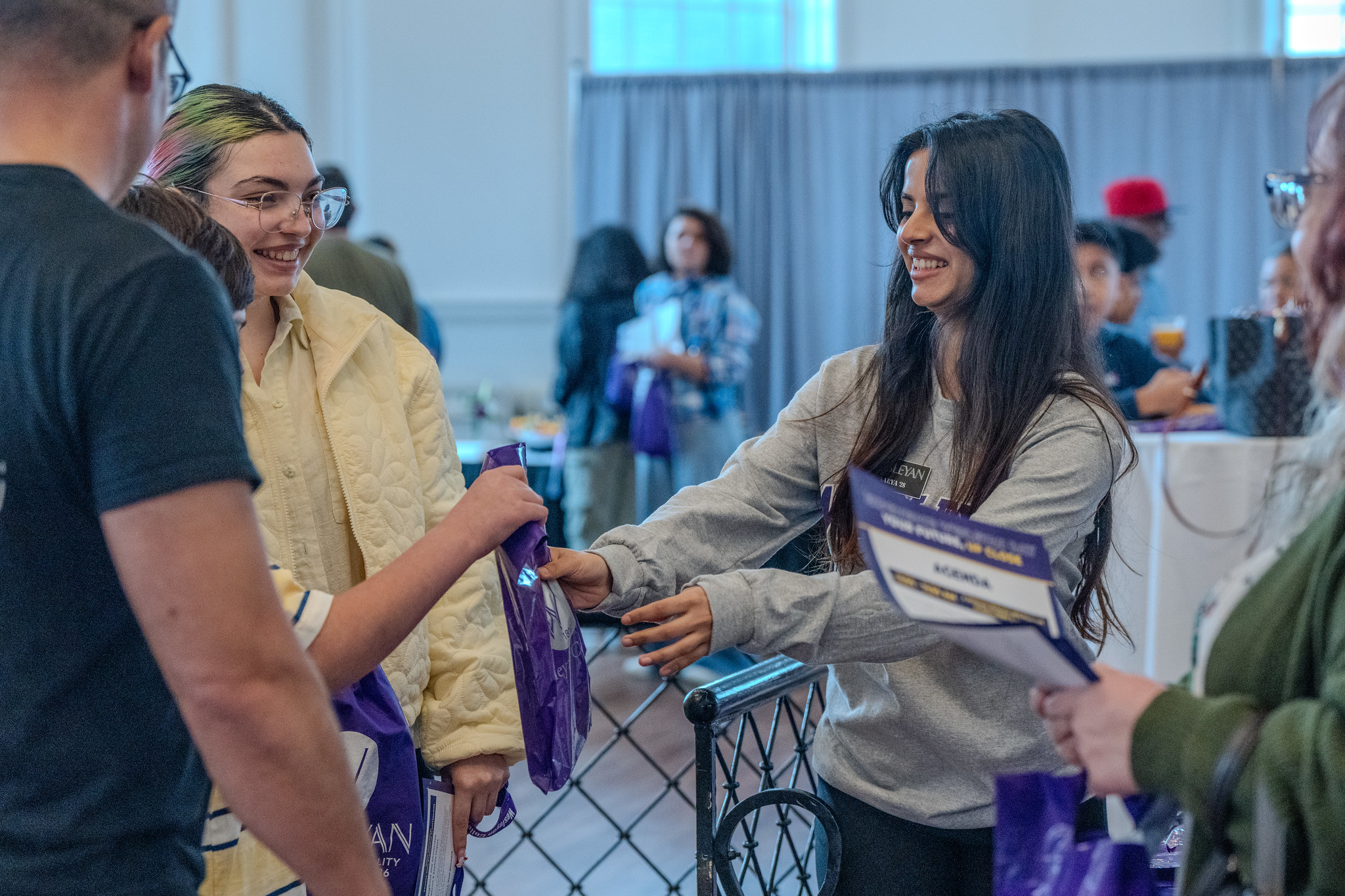 Two young women smiling and interacting at an indoor event, as one hands a purple bag to the other. They are surrounded by other attendees in a bright, casual setting with tables and informational materials visible in the background.