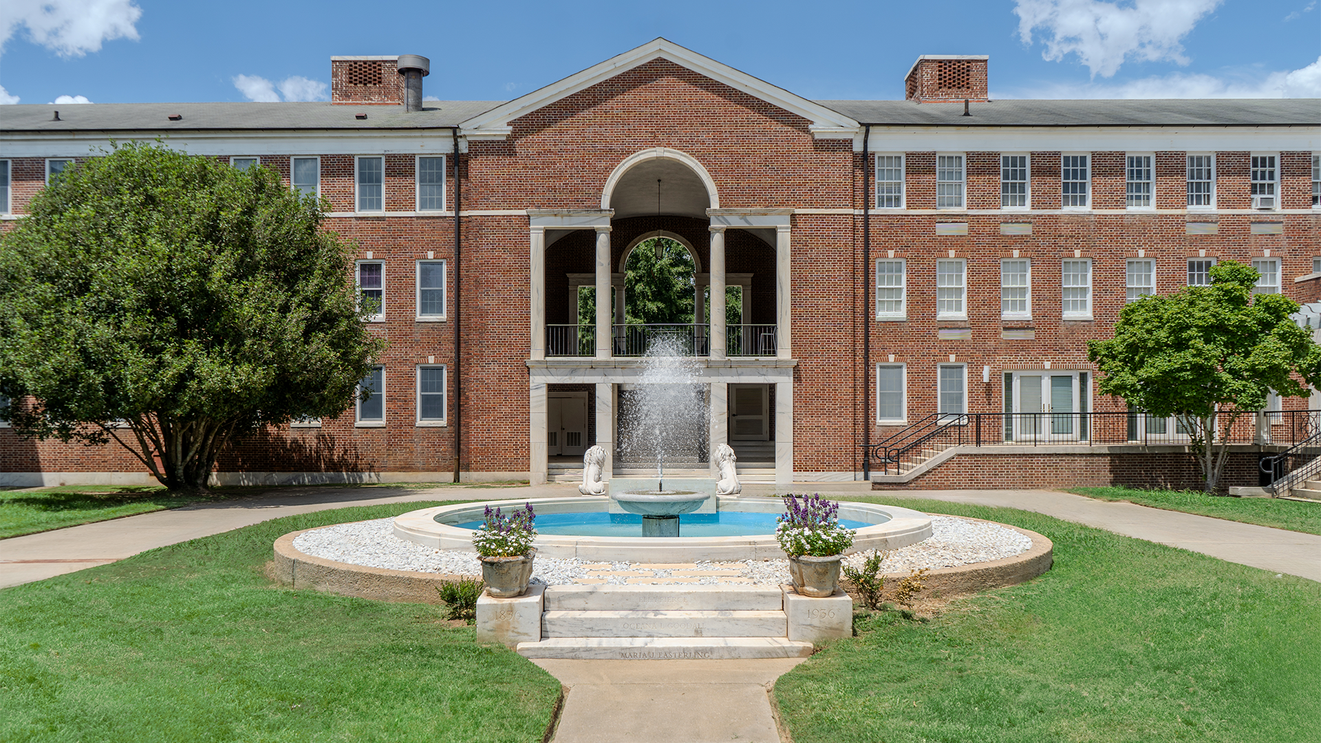 Large brick campus building with an arched central entrance and a fountain in the foreground, surrounded by green lawns, trees, and flower planters under a bright blue sky.