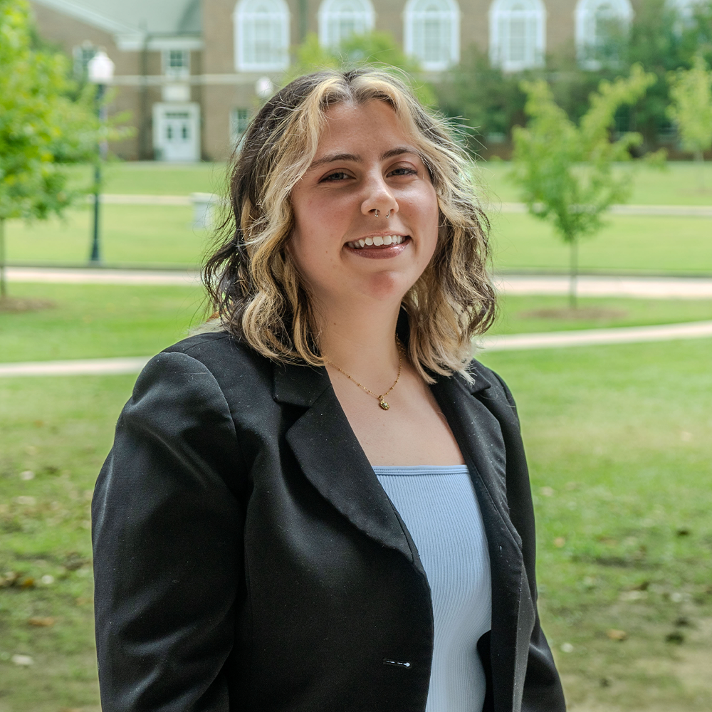Smiling young woman with short wavy hair featuring blonde highlights, standing outdoors on a campus lawn. She is wearing a black blazer over a light-colored top, with academic buildings and trees softly blurred in the background.