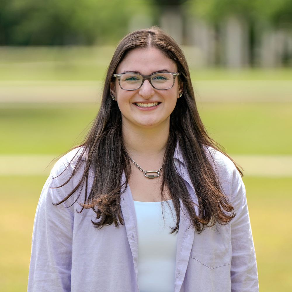 Smiling young woman with long dark hair and glasses standing outdoors on a grassy campus lawn. She is wearing a light button-up shirt over a white top and a silver chain necklace, with trees softly blurred in the background.