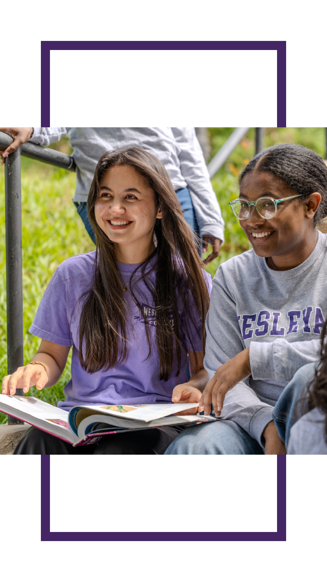 Two students sitting outdoors on campus steps, smiling and looking at an open book together while wearing Wesleyan apparel.