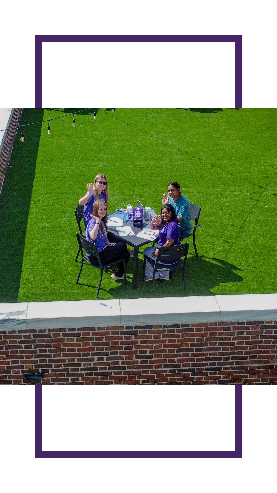 Four students seated around an outdoor table on a rooftop lawn, waving toward the camera during a casual study session.