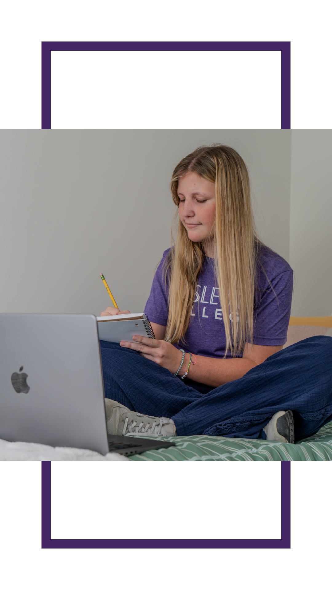Student sitting cross-legged on a bed while writing in a notebook beside an open laptop, studying in a dorm room.