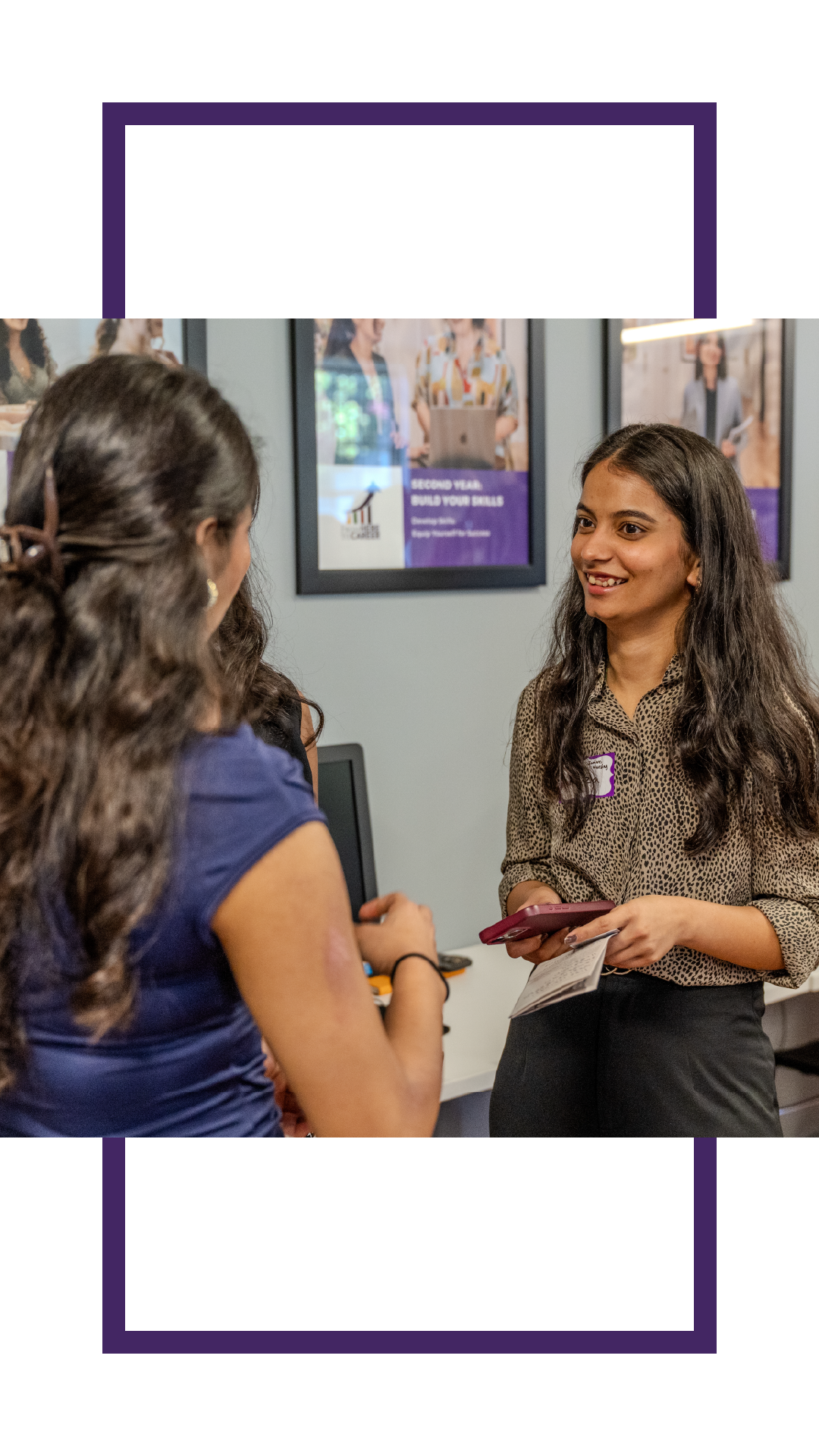 Student speaking with another student in an office setting, holding a phone and papers while smiling during a conversation. Career and professional development posters are displayed on the wall behind them.