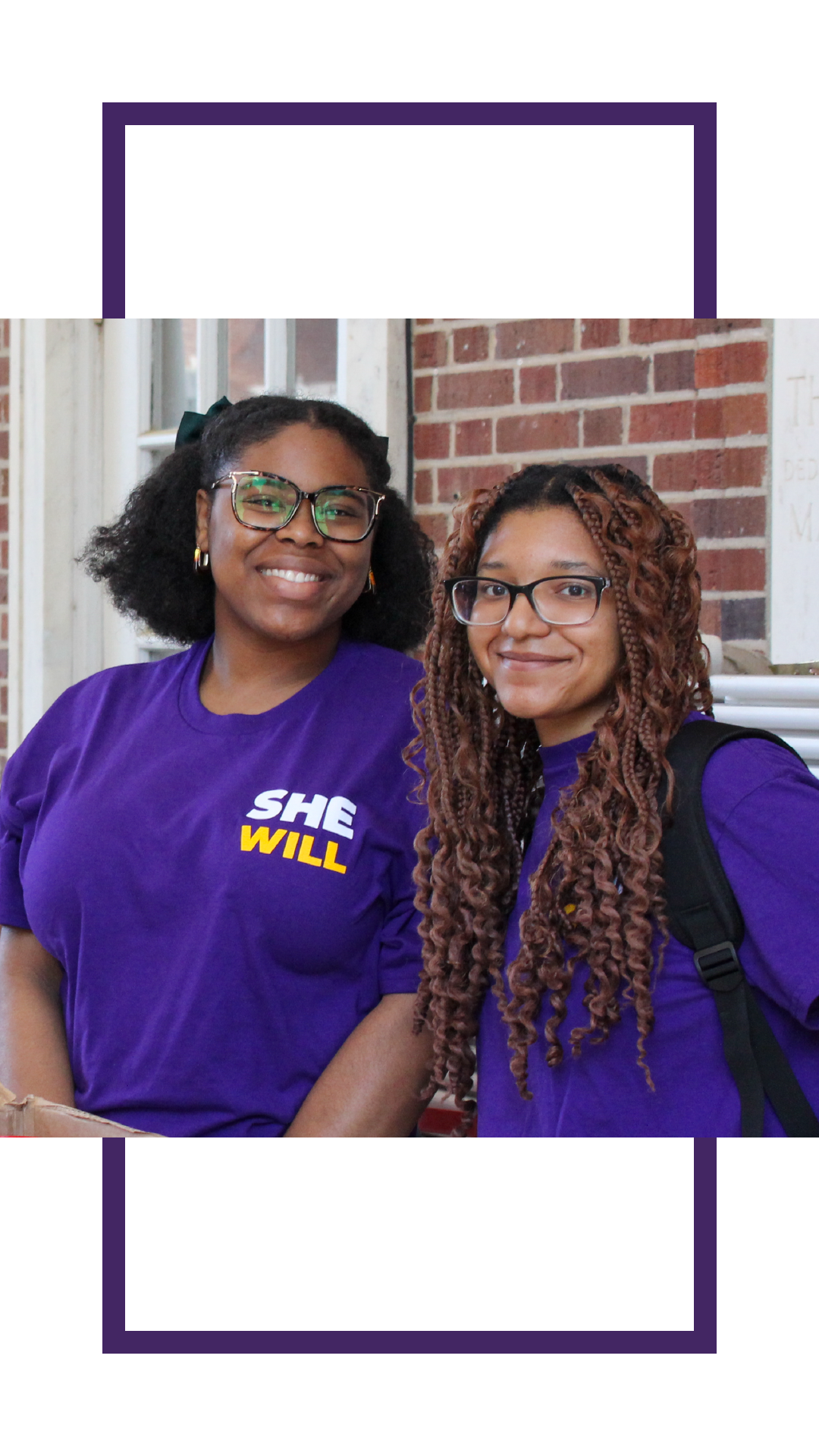 Two smiling students wearing purple “She Will” shirts stand together outside a brick campus building, one carrying a backpack.