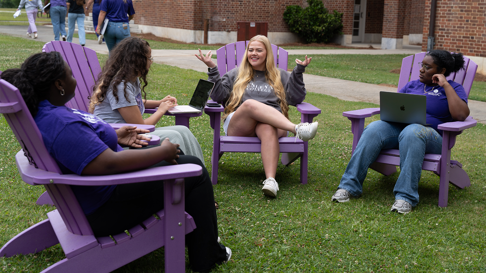 Group of students sitting in purple Adirondack chairs on a campus lawn, talking and using laptops outside a brick academic building. One student gestures while speaking as the others listen and interact in a relaxed outdoor study setting.