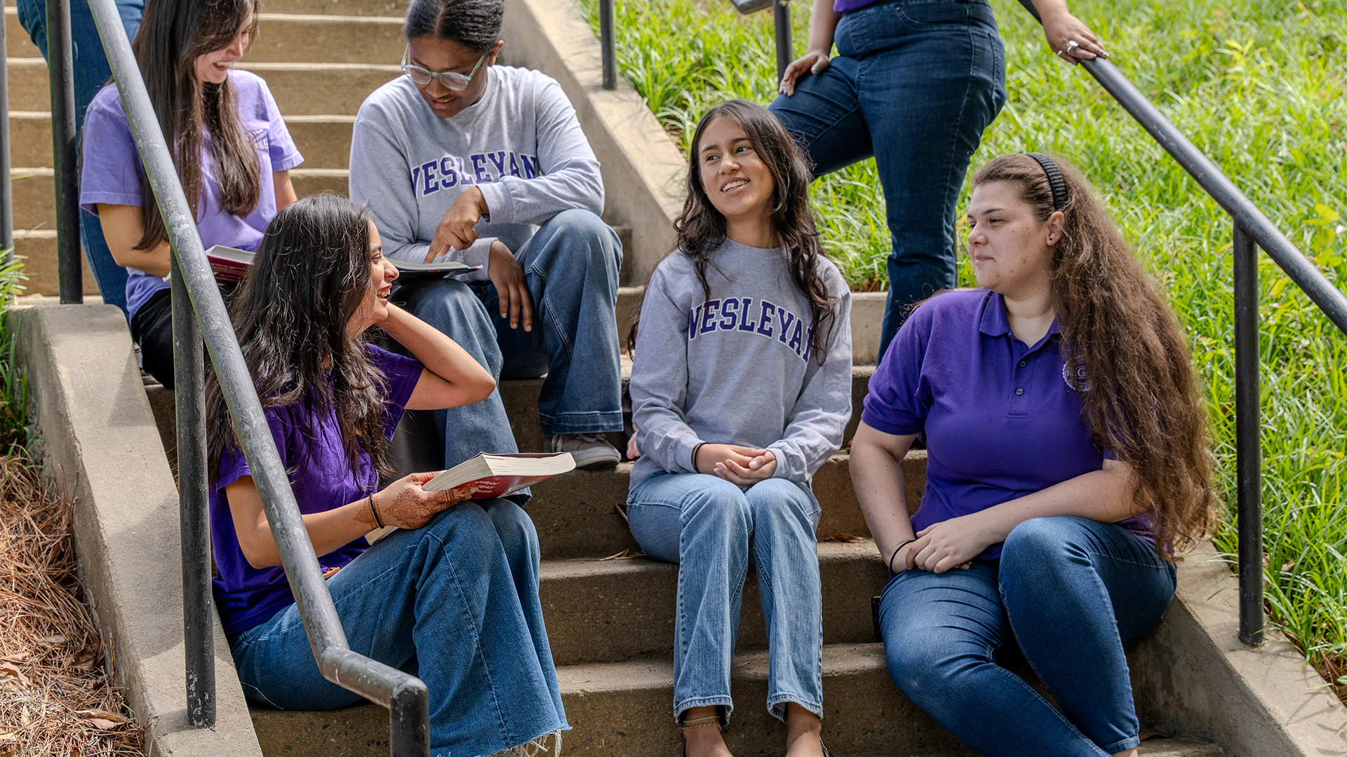 Group of students sitting together on outdoor campus steps, talking, reading, and smiling. Several wear Wesleyan shirts in purple and gray, creating a casual and collaborative study atmosphere surrounded by greenery.