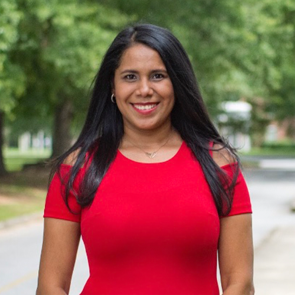 Smiling woman with long dark hair wearing a bright red dress, standing outdoors on a tree-lined campus walkway. The background is softly blurred with greenery and a road visible behind her.