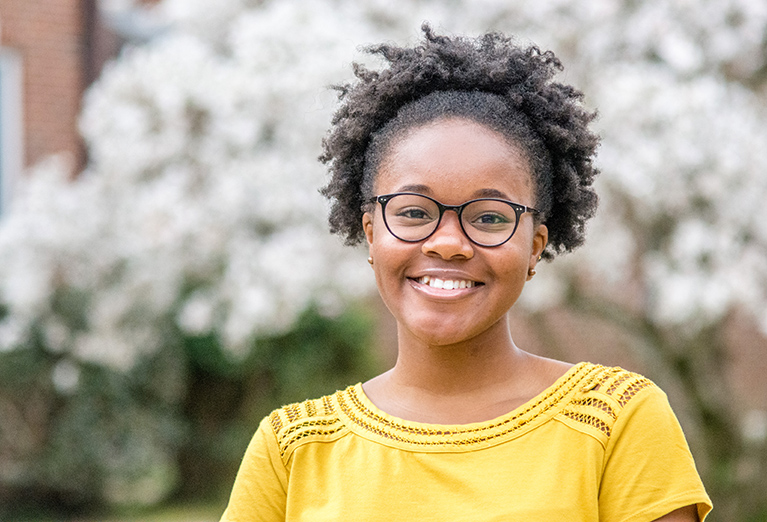 Student with glasses smiles outside in front of flowers.